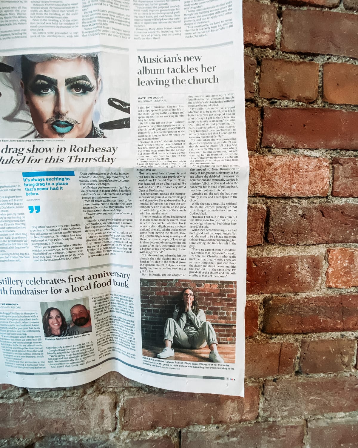 folded telegraph-journal article pinned against a brick wall — headline reads “musician’s new album tackles her leaving the church,” photo of tot sitting on the floor beneath.